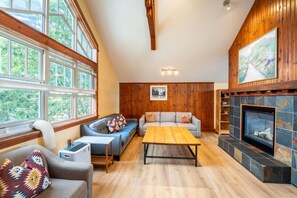 A cozy living room with a fireplace, a sofa, a coffee table, and large windows.