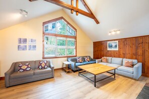 A spacious living room with a high ceiling, wooden beams, a large window, and a cozy seating area with sofas and a coffee table.