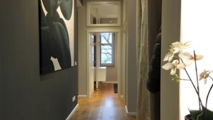 A hallway with a wooden floor, a door leading to a room, and a potted orchid on the right.