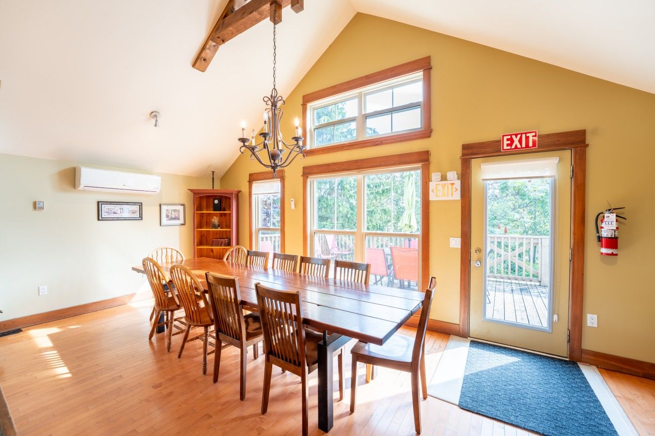 A dining area with a large wooden table, chairs, a chandelier, and a view through large windows.