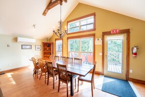 A dining area with a large wooden table, chairs, a chandelier, and a view through large windows.