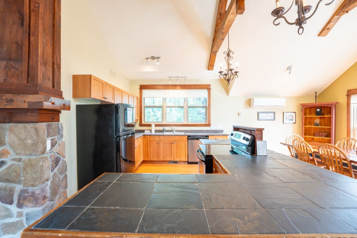 A kitchen with a stone wall, wooden cabinets, a black refrigerator, a stove, and a dining area with chairs.