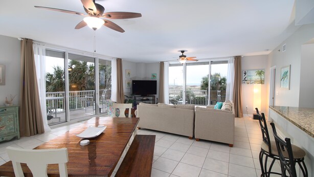 Un salón amplio con una mesa de comedor de madera, sofá beige, ventiladores de techo, ventanas grandes y un televisor.