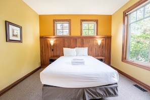 A hotel room with a large bed, wooden headboard, two windows, and a framed picture on the wall.