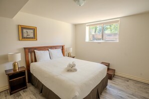 A bedroom with a large bed, two bedside tables, a nightstand, a framed picture on the wall, and a window with a view of trees.