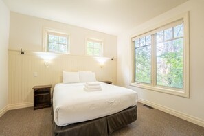 A bedroom with a large bed, two bedside tables, a nightstand, and a window with a view of trees. - The Prima at Blue View Chalets (The Blue Mountains)