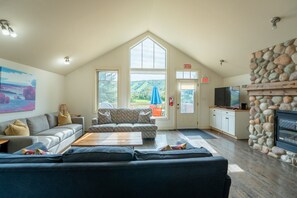 A spacious living room with a high ceiling, a stone fireplace, a large window, and a cozy seating area. - The Prima at Blue View Chalets (The Blue Mountains)