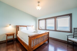 A bedroom with a wooden bed, a nightstand, a chair, and a large window with a view of the sea. - The Aqua Villa at Blue View Chalets (The Blue Mountains)