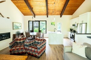 A spacious living area with a dining table, two sofas, a kitchen with modern appliances, and a large window offering a view of greenery.