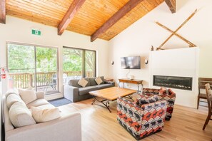 A spacious living area with a wooden ceiling, a fireplace, a television, and a balcony with outdoor seating.