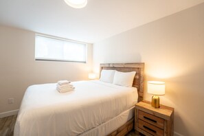 A bedroom with a bed, a nightstand with a lamp, a window, and a wall socket. - The 44L at Blue View Chalets (The Blue Mountains)