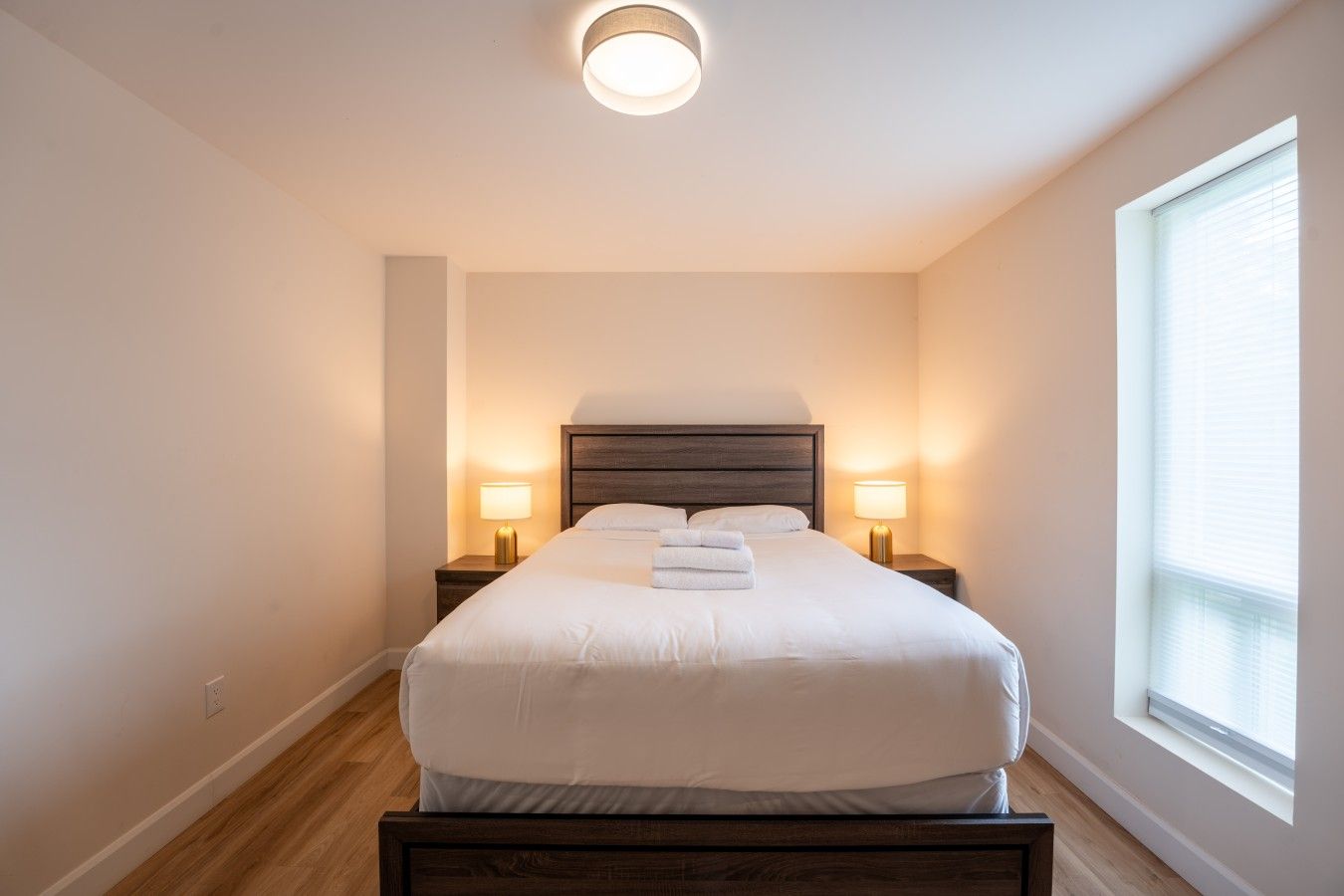 A neatly made bed in a room with a large window, two bedside lamps, and a ceiling light.