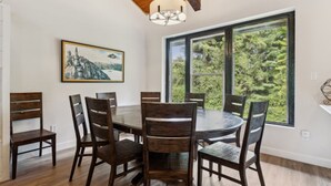 A dining area with a round table and six chairs, large windows with a view of greenery, and a ceiling fan. - The 44L at Blue View Chalets (The Blue Mountains)