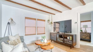 A living room with a sofa, round wooden coffee table, and a TV. There are windows with blinds, a wooden cabinet, and a hanging light fixture.