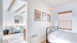 A bedroom with a bed, a window with blinds, a radiator, a sofa, and a wall with framed pictures.