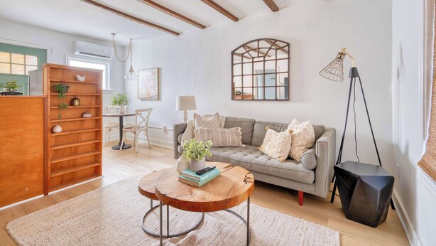 A cozy living room with a wooden bookshelf, a round coffee table with books and a plant, a grey sofa with cushions, and a tall floor lamp.