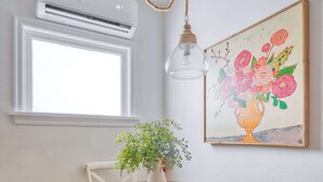 A dining area with a round wooden table, two white chairs, a wicker chair, a vase with green plants, a hanging light fixture, an air conditioning unit, and a painting of flowers on the wall.