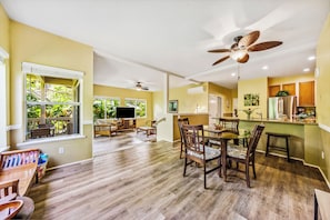 Relax in this inviting living area with a dining table for four, a flat-screen TV, and a large window offering a view of lush greenery.