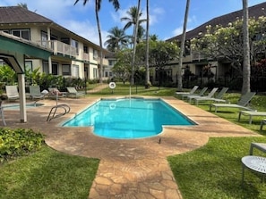 Relax by the pool surrounded by lush greenery and palm trees, with lounge chairs and a table for a peaceful retreat.