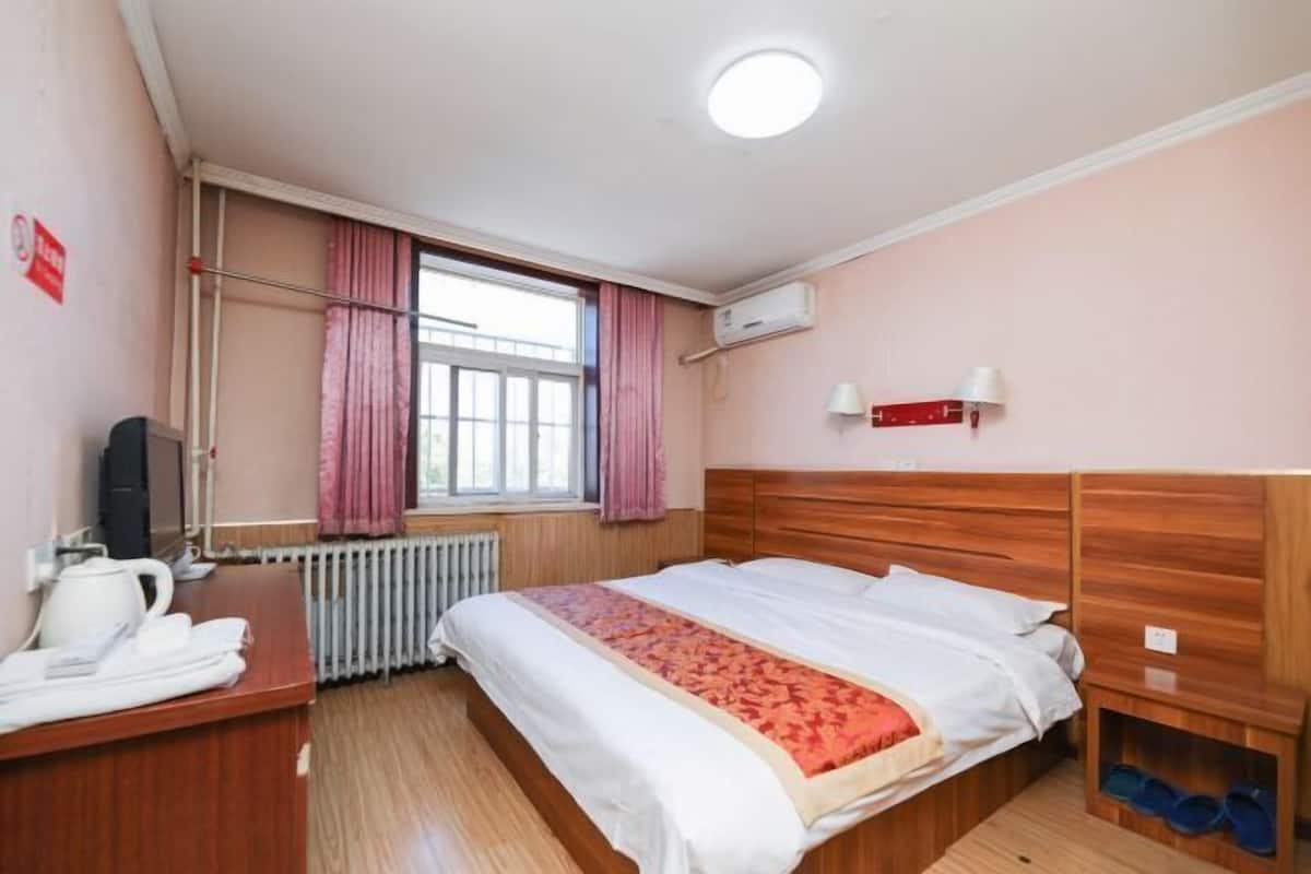 A hotel room with a wooden bed, a bedside table with a kettle, a radiator, a window with curtains, and an air conditioning unit.