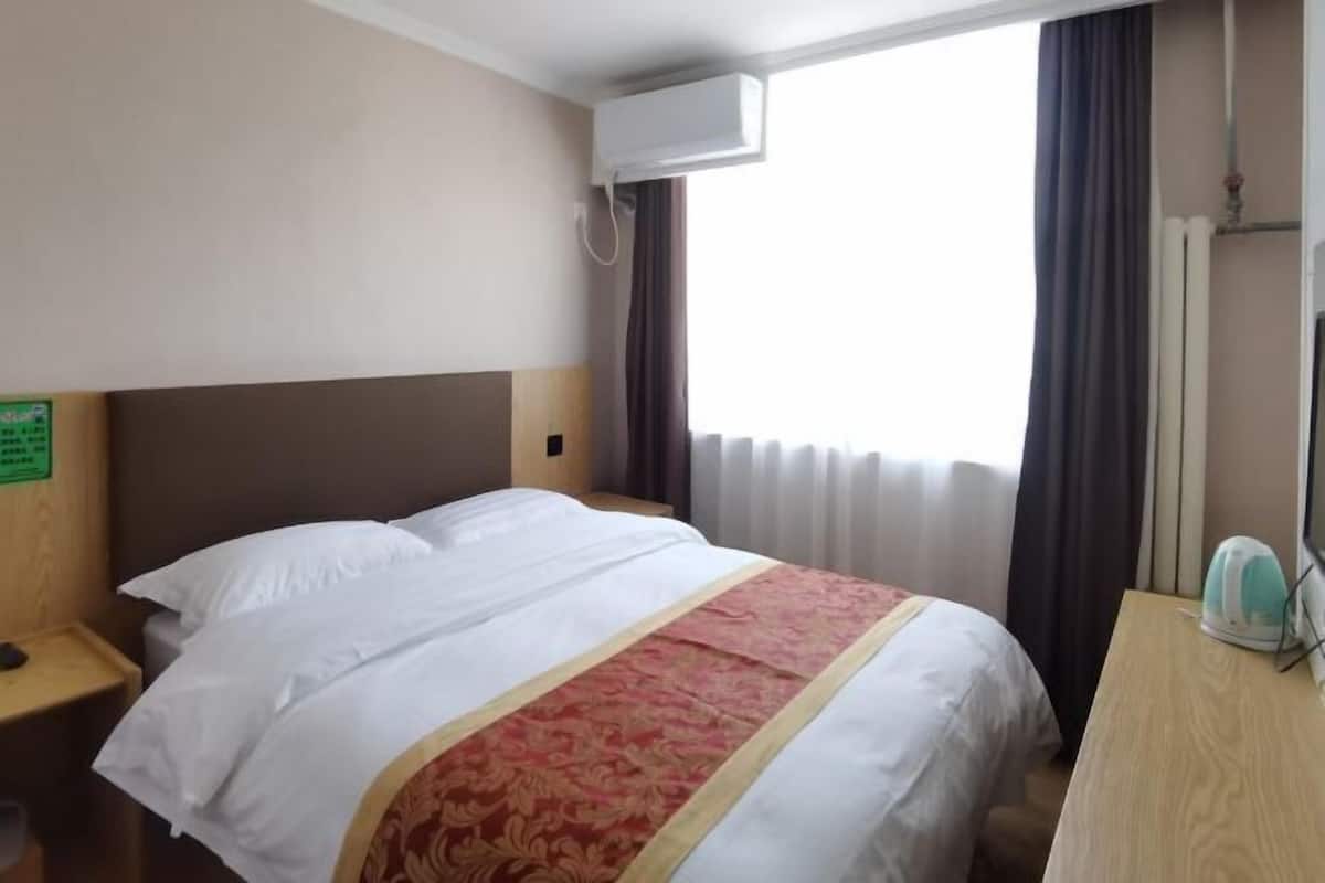 A hotel room with a bed, a nightstand, a desk, an air conditioning unit, and a window with curtains.