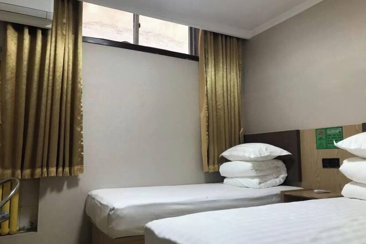 A hotel room with two single beds, a window with curtains, a wooden headboard, and a small bedside table.