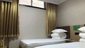 A hotel room with two single beds, a window with curtains, a wooden headboard, and a small bedside table.