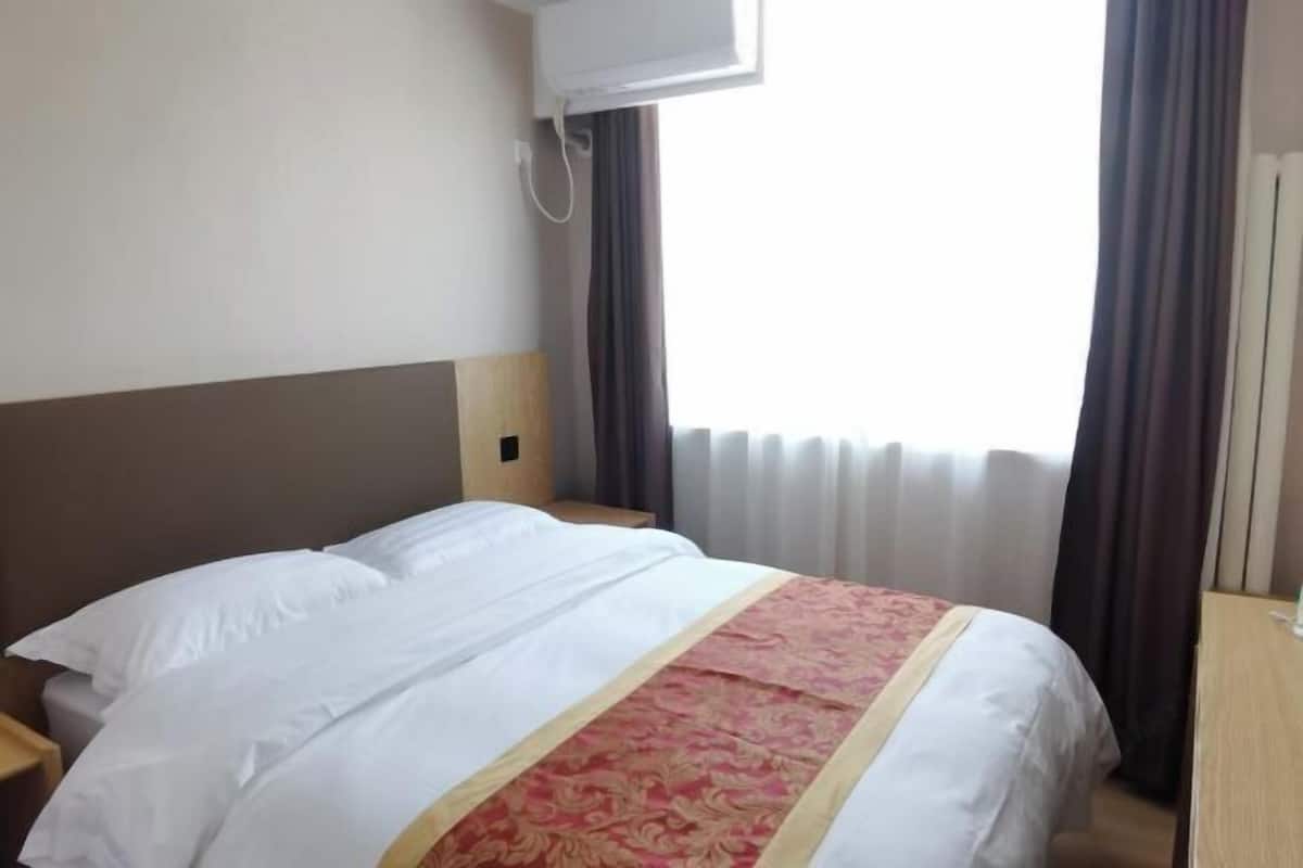 A hotel room with a bed, a nightstand, a window with curtains, and an air conditioning unit.