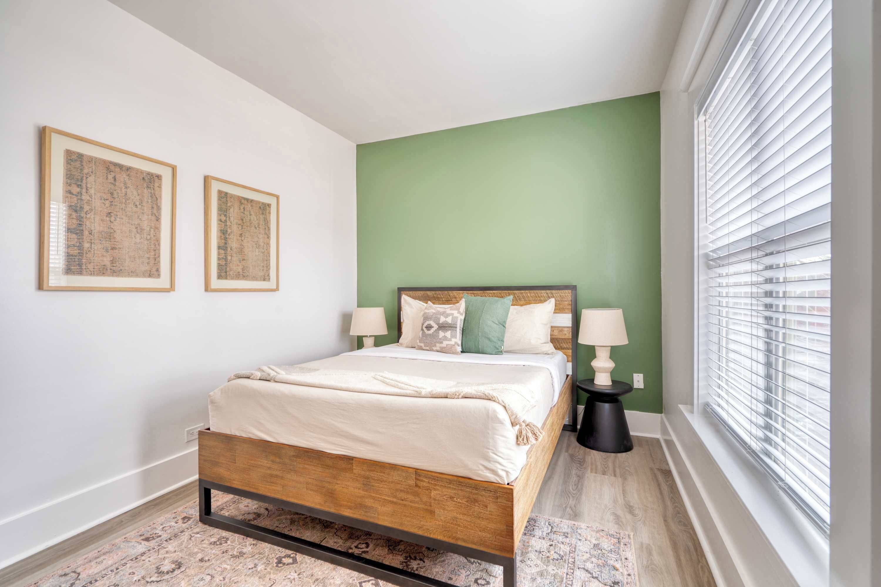 A bedroom with a wooden bed, green walls, two framed pictures, a nightstand with a lamp, and a window with blinds.
