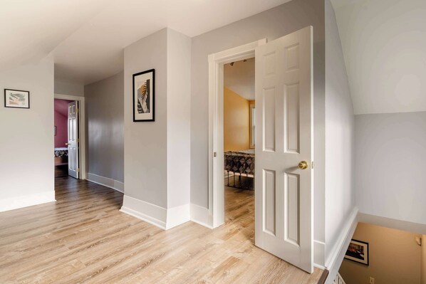A hallway with wooden flooring, white walls, and a door leading to a room with a bed. - Remodeled 3BR - Walk to Coffee, Bars & More (Portland)