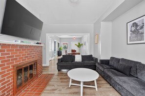A modern living room with a fireplace, grey sofas, a white coffee table, and a flat-screen TV mounted above the fireplace. - Remodeled 3BR - Walk to Coffee, Bars & More (Portland)