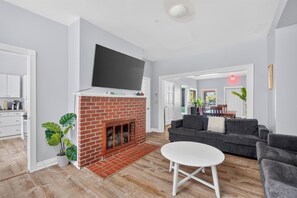 A modern living room with a brick fireplace, a flat-screen TV mounted above it, a grey sofa, and a white coffee table. - Remodeled 3BR - Walk to Coffee, Bars & More (Portland)