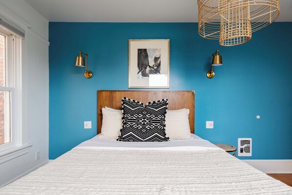 A bedroom with a blue wall, a wooden headboard, a bed with white bedding, two black and white patterned pillows, a framed picture above the bed, and two wall-mounted lamps.