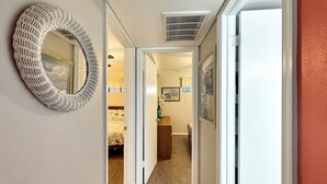 Un pasillo con un espejo tejido a la izquierda, una puerta que da a una habitación con cama y ventana, y un cuadro en la pared.