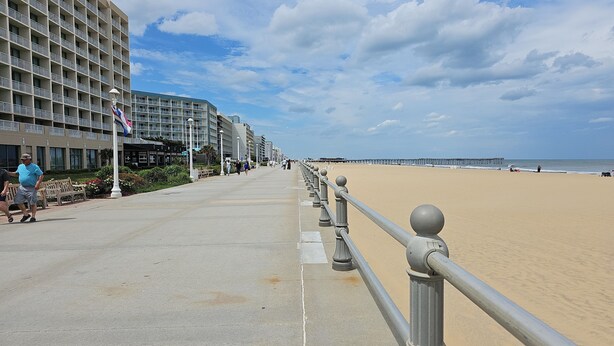 Un paseo marítimo frente a la playa con un camino de hormigón, un muelle a lo lejos y un edificio de hotel con balcones.