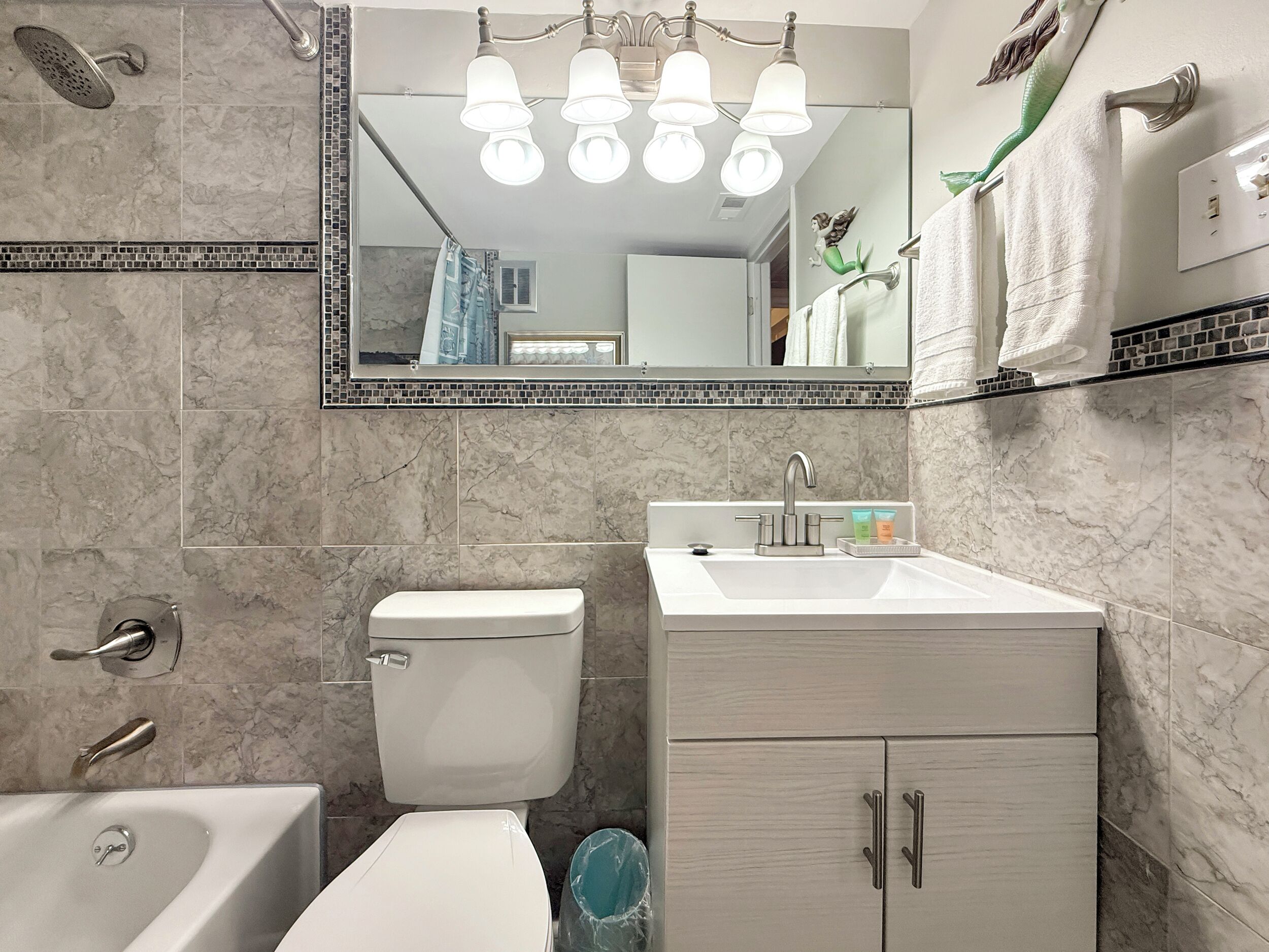 Un baño con inodoro, lavamanos y bañera. Hay un espejo sobre el lavamanos, y una ducha sobre la bañera.