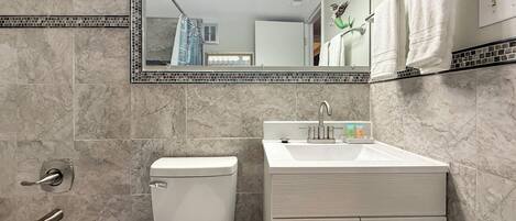 Un baño con inodoro, lavamanos y bañera. Hay un espejo sobre el lavamanos, y una ducha sobre la bañera.