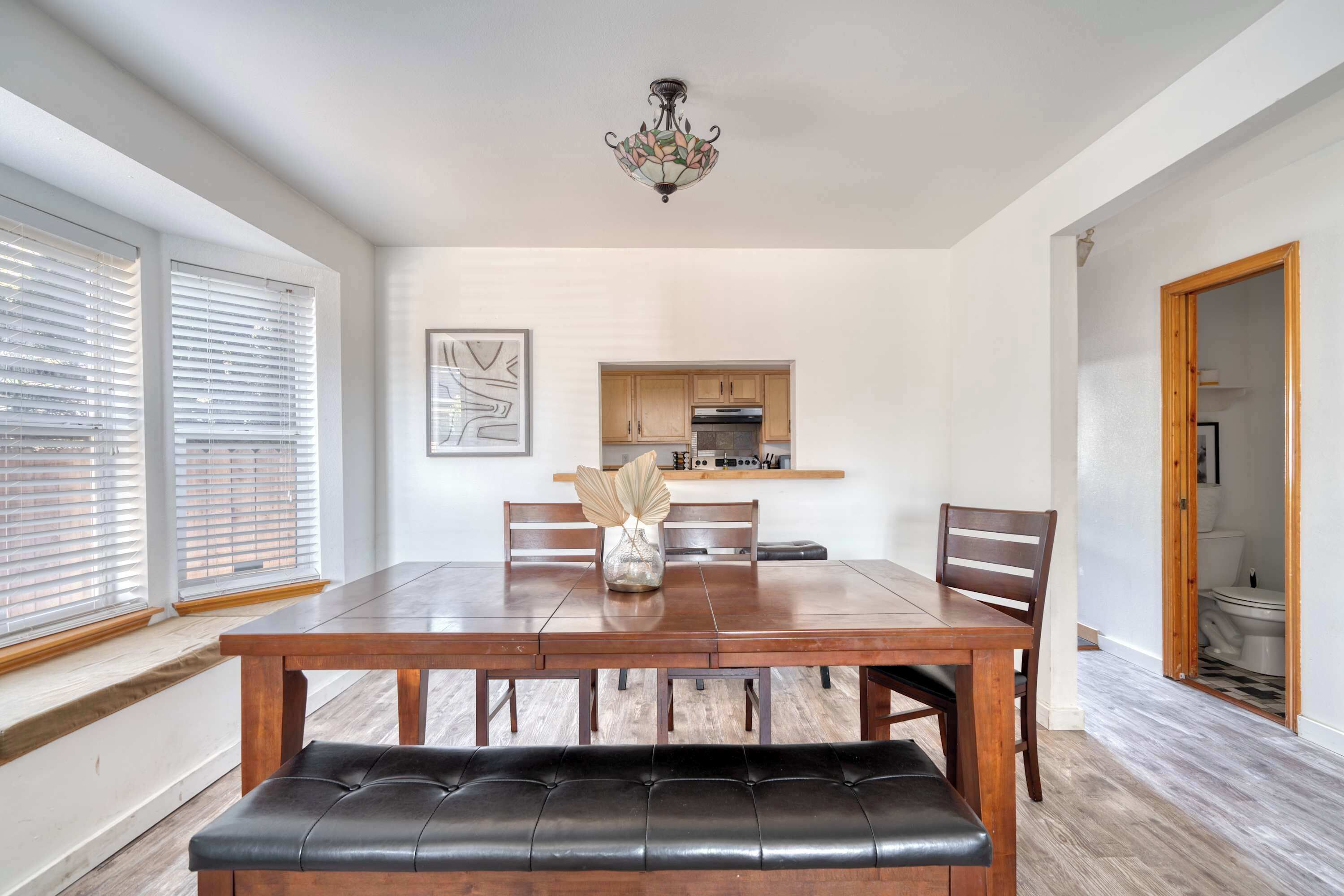 Un comedor con una mesa de madera, sillas y un banco. Se observa una cocina al fondo con armarios de madera y una encimera.