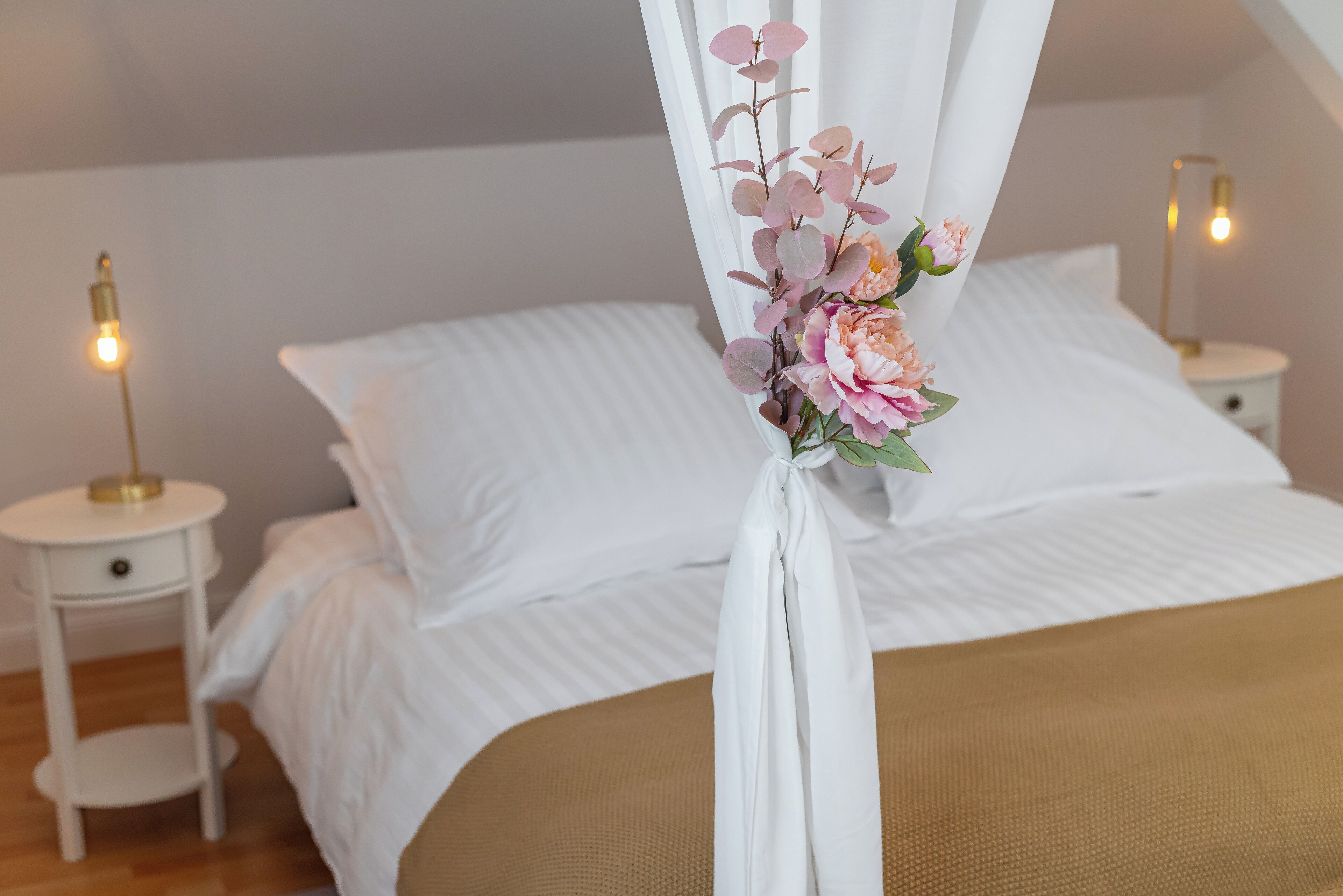 A neatly made bed with white linens, a canopy with floral decorations, and bedside tables with lamps.