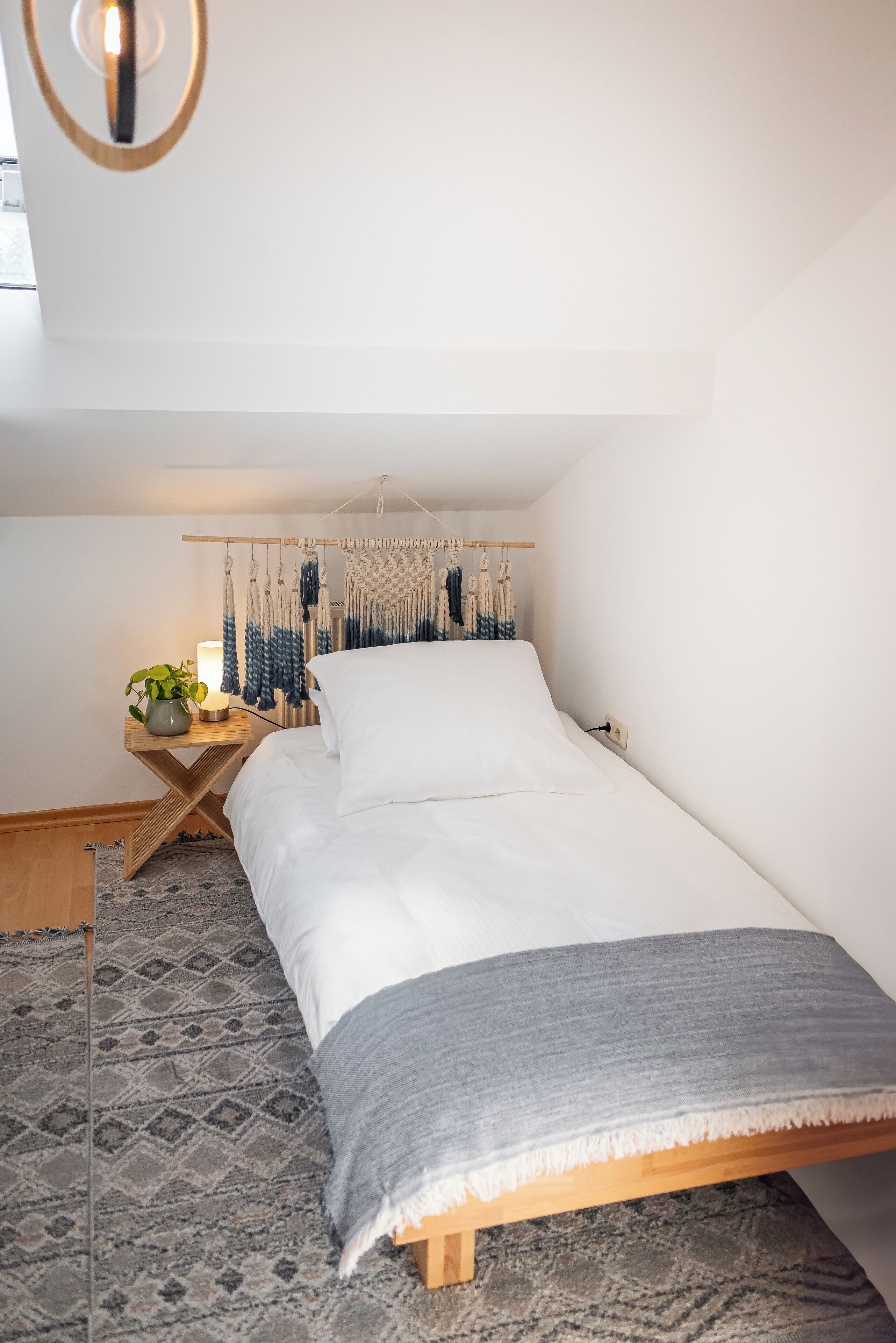 A bedroom with a bed, bedside table, lamp, and wall hanging.