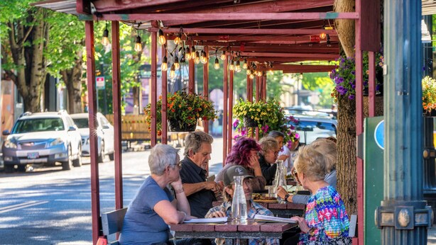 Un espace extérieur aménagé avec des tables et des chaises, protégé par un toit, et entouré d’arbres et de voitures garées.