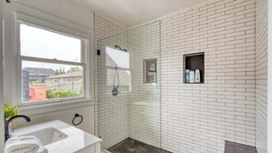 A bathroom with a white-tiled shower, a glass-enclosed shower area, a white sink with a marble countertop, and a window providing a view of buildings outside.