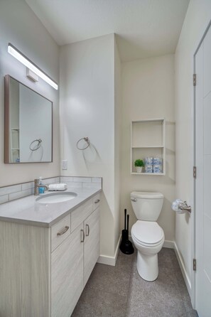 A modern bathroom with a white sink, mirror, and toilet. There is a shelf above the toilet with toiletries and a small plant.