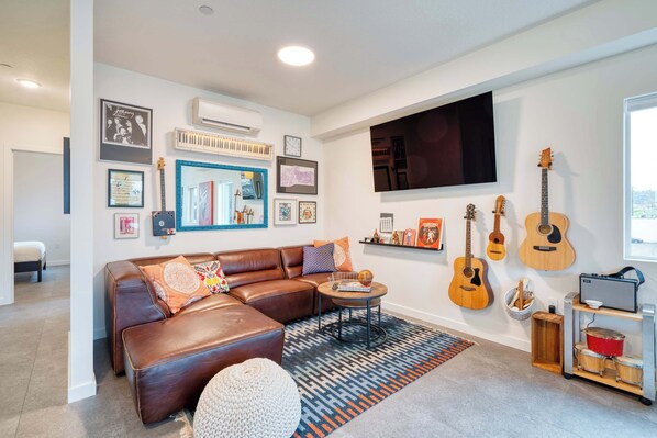 A modern living room with a brown leather sofa, a coffee table, and a TV mounted on the wall. There are several guitars and a drum set on a shelf. - Luxury Penthouse - Rooftop Deck - 3BR - AC (Portland)