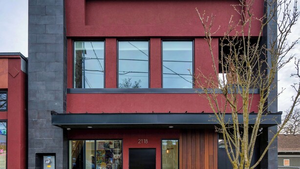 A modern two-story building with a red facade, large windows, and a small tree in the foreground.