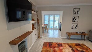 A living room with a fireplace, a TV mounted on the wall, a dining area with a table and chairs, and a large colorful rug. - 3 Bed-sleeps 5-garden-parking-pets (Carlisle)
