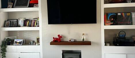 A living room with a fireplace, a flat-screen TV mounted on the wall, and shelves with various items.