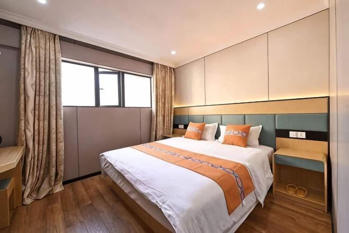 A hotel room with a large bed, a desk, a window with curtains, and a headboard with built-in lighting.