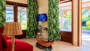 A room with a glass-top table, a red sofa, a television mounted on the wall, and curtains with a leaf pattern. - Charming villa in Pereybère and walking distance to the beach with private pool. (Grand Baie)