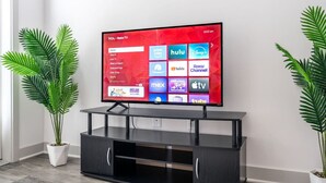 A TCL Roku TV mounted on a black entertainment center with a red interface displaying various streaming apps. - Stylish Stay | Near Everything! (Whitestown)